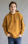 A man wearing a mustard yellow hoodie with a blue shield logo on the left chest and blue graphics on the right sleeve, paired with blue jeans, standing against a neutral gray background.