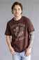 A man wearing a brown graphic t-shirt with 'Ariat Boots Tail Worn' printed in distressed white lettering and a cowboy skull design, paired with light blue jeans against a gray studio background.
