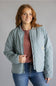 A woman wearing a light sage green quilted jacket by Carhartt, layered over a dusty rose top and blue jeans, standing against a solid gray studio background.
