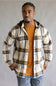 A man wearing a cream-colored hooded plaid overshirt with navy and mustard yellow stripes, layered over an orange t-shirt and blue jeans, standing against a neutral gray background.