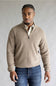 A man wearing a light beige ribbed quarter-zip pullover sweater over a cream collared shirt and blue jeans, standing against a neutral gray studio background.