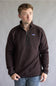 A man wearing a dark brown Patagonia quarter-zip fleece pullover sweater and blue jeans, standing against a neutral gray studio background.