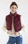 A smiling woman wearing a burgundy quilted puffer vest over a cream cable-knit sweater and blue jeans, photographed against a plain white studio background.