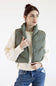 A woman wearing an olive green puffer vest over a cream cable-knit sweater and blue jeans, standing against a plain white studio background.