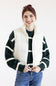 A smiling woman wearing a cream quilted puffer vest over a dark green and cream striped knit sweater, paired with blue jeans, against a plain white studio background.