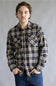 A man wearing a long-sleeve plaid button-up shirt in brown, beige, and white, paired with blue jeans, standing against a neutral gray studio background.