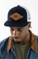 A man wearing a navy blue and white trucker hat with a brown diamond-shaped patch on the front, styled with a denim jacket and olive green t-shirt against a plain white background.