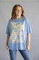 A woman modeling a light blue oversized graphic t-shirt with a large yellow vintage-style print on the front, paired with blue jeans, against a solid gray studio background.