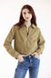 A woman with red hair wearing a cropped tan zip-up jacket and blue jeans, standing against a plain white studio background.
