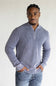 A man wearing a ribbed long-sleeve half-zip sweater in a dusty periwinkle blue color, paired with blue jeans, standing against a plain light gray background.