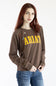 A woman with red hair wearing a brown crewneck sweatshirt with 'ARIAT' printed in yellow across the chest, paired with blue jeans, against a plain white background.