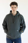 A man wearing a dark green pullover hoodie with drawstrings and a kangaroo pocket, paired with blue jeans, standing against a plain white background.