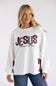 A woman wearing a white long-sleeve sweatshirt with 'JESUS is the reason for the season' printed in red and black plaid and white on the front, paired with blue jeans, against a light gray studio background.
