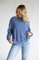A woman wearing a dusty blue long-sleeve mock neck sweatshirt and light wash blue jeans, standing against a plain light gray studio background.