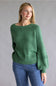 A woman modeling a green textured knit sweater with a boat neckline and blue jeans, standing against a solid gray studio background.