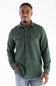 A man wearing a dark green long-sleeve button-up shirt with two chest pockets and light blue jeans, standing against a plain white background.