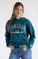 A woman wearing a teal green hoodie with 'BOOK CLUB Since 1989' printed in pink and white on the front, paired with blue jeans, against a plain white background.