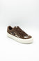 A low-top sneaker in brown suede and canvas with a cow-print pattern on the side, brown laces, and a thick white rubber sole, shown against a plain white background.