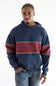 A man wearing a navy blue hoodie with a wide burgundy horizontal stripe across the chest and thin gold trim, paired with blue jeans, against a plain white background.