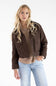 A blonde woman modeling a dark brown cropped utility jacket over a beige ribbed sweater and light blue jeans, photographed against a clean white studio background.