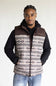A man wearing a brown and beige patterned zip-up vest over a black hoodie and blue jeans, standing against a plain light background. The vest features a geometric Southwestern-style pattern and a small 'Ariat' logo on the left chest.