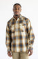 A man wearing a Carhartt long-sleeve plaid button-up shirt in beige, brown, and blue tones, paired with khaki pants, photographed against a plain white studio background.