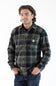 A man wearing a long-sleeve plaid button-up shirt in dark green, black, and gray, paired with blue jeans, standing against a plain white background.
