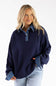 A woman wearing an oversized navy knit sweater with a denim collar and matching denim cuffs, paired with light blue jeans, against a plain white background.
