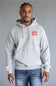 A man wearing a light gray heather pullover hoodie with a red 'The North Face' logo patch on the left chest, paired with blue jeans, against a solid gray studio background.
