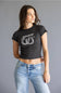 A woman modeling a black short-sleeve crop top with a white Billabong logo graphic on the chest, paired with light blue jeans, against a solid gray studio background.