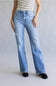 A person wearing light-wash blue distressed flare jeans with frayed hems, paired with a striped top and black shoes, standing against a plain light gray studio background.