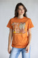 A woman modeling an orange graphic t-shirt with a 'Fall' theme featuring cowboy boots, pumpkins, and the text 'simply southern.' paired with light blue jeans, against a plain light gray background.
