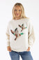 A woman wearing a cream-colored knit sweater with a large brown and green bird motif across the chest, paired with blue jeans, against a plain white background.