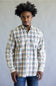 A man wearing a long-sleeve button-down plaid shirt in beige, blue, and white, paired with blue jeans, standing against a plain gray studio background.