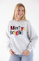 A person wearing a light heather gray crewneck sweatshirt with the words 'Merry & Bright' printed across the chest in multicolored textured letters.