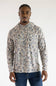 A man wearing a long-sleeve hooded shirt with an abstract organic camouflage pattern in beige, brown, and gray, paired with blue jeans, against a plain white studio background.