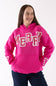 A woman wearing a bright pink pullover hoodie with 'MERRY' written across the chest in large white letters outlined in red, paired with dark blue jeans, against a plain white background.