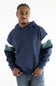 A man wearing a navy blue hoodie with white and green color-blocked stripes on the sleeves, paired with blue jeans, photographed against a plain white studio background.