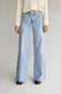 A model from the waist down wearing light-wash blue wide-leg jeans with a drawstring waist, paired with a cream ribbed sweater and brown shoes, against a light gray studio background.