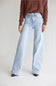 A model wearing light-wash wide-leg denim jeans, a brown ribbed knit sweater, and black pointed-toe heels, standing against a neutral light gray studio background.
