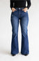 A person wearing dark blue high-rise flare jeans with distressed details and a black long-sleeve top, standing against a plain white studio background.