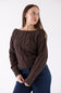 A woman wearing a dark brown cable-knit off-the-shoulder sweater and dark blue jeans, standing against a plain white studio background.
