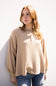 A woman with long light brown hair wearing a beige waffle-knit long-sleeve sweater and blue jeans, standing against a white wall.