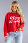 A smiling blonde woman wearing a bright red knit sweater with 'Merry Bright' in white script across the front, paired with light blue jeans, against a solid gray studio background.