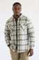 A man wearing a long-sleeve button-up plaid overshirt in beige and olive green, paired with khaki pants, standing against a plain white studio background.