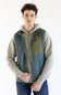 A man wearing a sleeveless zip-front vest over a light gray hoodie and blue jeans, standing against a plain white studio background.