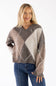 Front view of a smiling woman with long blonde hair wearing a brown Miracle argyle V-neck sweater featuring a soft fuzzy knit fabric and an all-over diamond pattern, paired with dark denim jeans.
