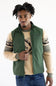 A man wearing a green zip-up vest over a beige and gray patterned long-sleeve shirt, paired with blue jeans, against a plain white background.