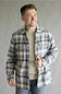 A smiling man wearing a long-sleeve plaid button-up shirt in blue, gray, and beige over a light beige crewneck sweater and blue jeans, standing against a neutral gray studio background.