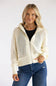 A woman modeling a cream open-knit zip-up hoodie jacket over a black top and blue jeans, photographed against a plain light gray studio background.